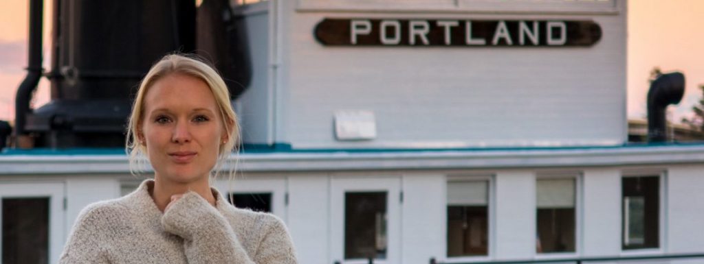 Courtenay stands on a dock in front of a large blue and white boat in Portalnd during sunset. She wears a beige cardigan and rests on arm across her stomach with her chin perched on the knuckles of her opposite hand.